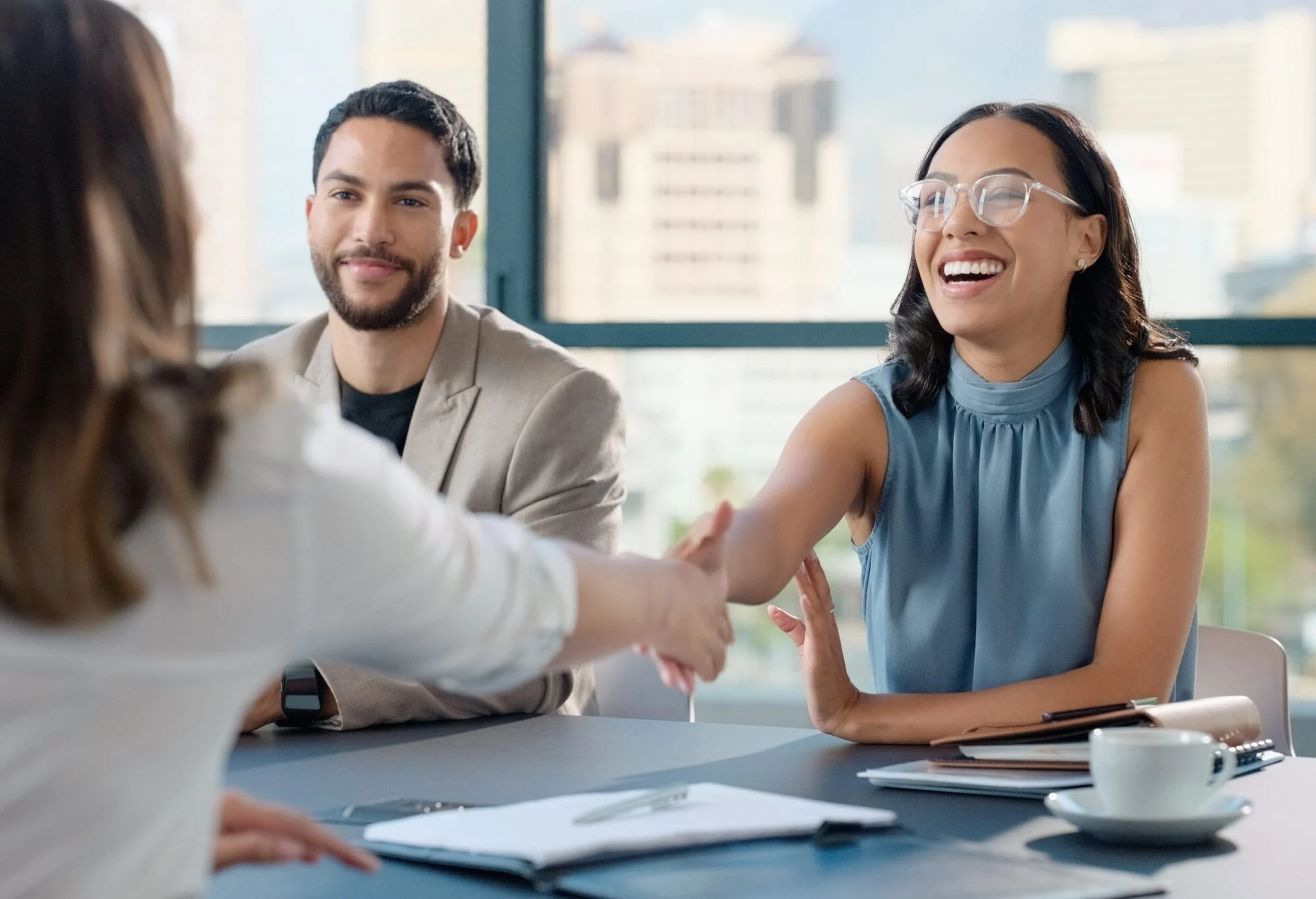 Smiling woman in glasses shakes hands with another person across a table during a meeting.